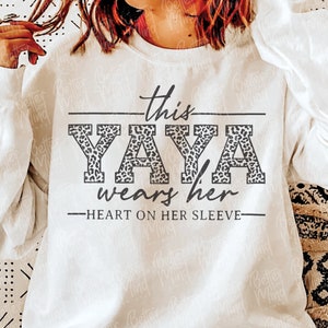 Puede incluir: Sudadera blanca con el texto "this YAYA wears her heart on her sleeve" en letras negras con un diseño de estampado de leopardo detrás de la palabra "YAYA".