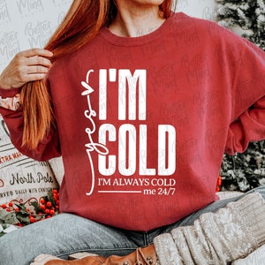 May include: A maroon sweatshirt with the text "I'M COLD" in white, along with the phrase "I'M ALWAYS COLD me 24/7". The image also includes a pillow with the text "HOT SINCE" and a Christmas theme.
