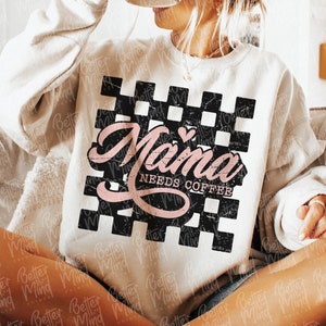 Op de afbeelding: Een wit sweatshirt met een zwart-wit geruit ontwerp en de tekst "Mama Needs Coffee" in roze letters met een klein roze hart.