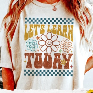 May include: A white t-shirt with a retro-style graphic that says "Let's Learn Today" in colorful letters with flowers and stars. The graphic is surrounded by a checkered border.