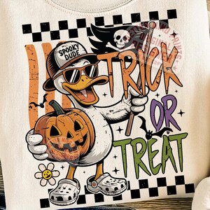 May include: Cream-colored sweatshirt with a Halloween-themed graphic. The design features a cartoon duck wearing sunglasses and a hat, holding a carved pumpkin and a flag that says "Trick or Treat". The duck is wearing Crocs and the text "Spooky Dude".