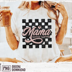 Op de afbeelding: Een wit T-shirt met een zwart-wit geruite achtergrond. De tekst "Mama Needs Coffee" is geschreven in roze schrift met een klein roze hartje boven het woord "Mama".