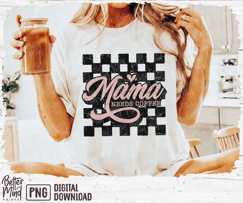 Op de afbeelding: Een wit T-shirt met een zwart-wit geruite achtergrond. De tekst "Mama Needs Coffee" is geschreven in roze schrift met een klein roze hartje boven het woord "Mama".