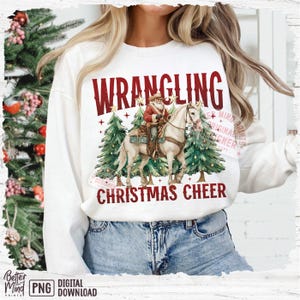 May include: White sweatshirt with a graphic featuring Santa Claus riding a horse between two Christmas trees. The text "WRANGLING CHRISTMAS CHEER" is printed in red. The design has a watercolor style.