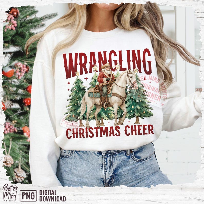 May include: White sweatshirt with a graphic featuring Santa Claus riding a horse between two Christmas trees. The text "WRANGLING CHRISTMAS CHEER" is printed in red. The design has a watercolor style.