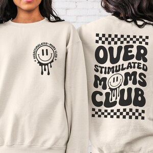 Pode incluir: Uma camisola de cor creme com o texto "Over Stimulated Moms Club" em preto nas costas. A frente da camisola tem uma cara sorridente preta a pingar com o texto "Over Stimulated Moms Club" &agrave; volta.