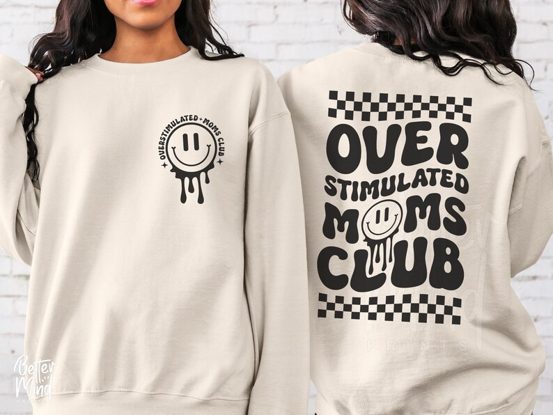 Pode incluir: Uma camisola de cor creme com o texto "Over Stimulated Moms Club" em preto nas costas. A frente da camisola tem uma cara sorridente preta a pingar com o texto "Over Stimulated Moms Club" &agrave; volta.