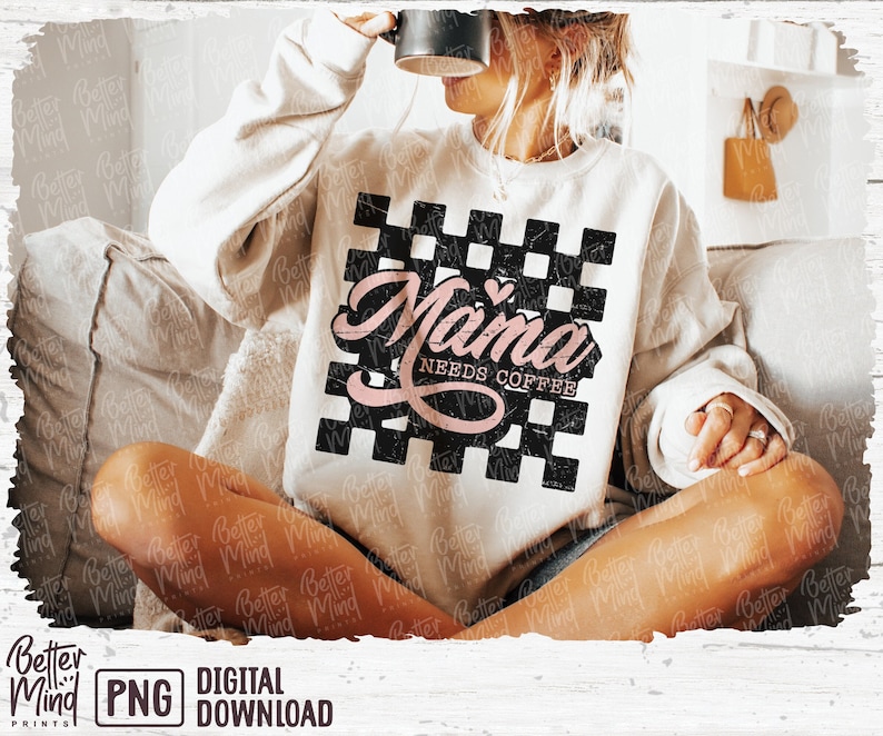Op de afbeelding: Een wit sweatshirt met een zwart-wit geruit ontwerp en de tekst "Mama Needs Coffee" in roze letters met een klein roze hart.