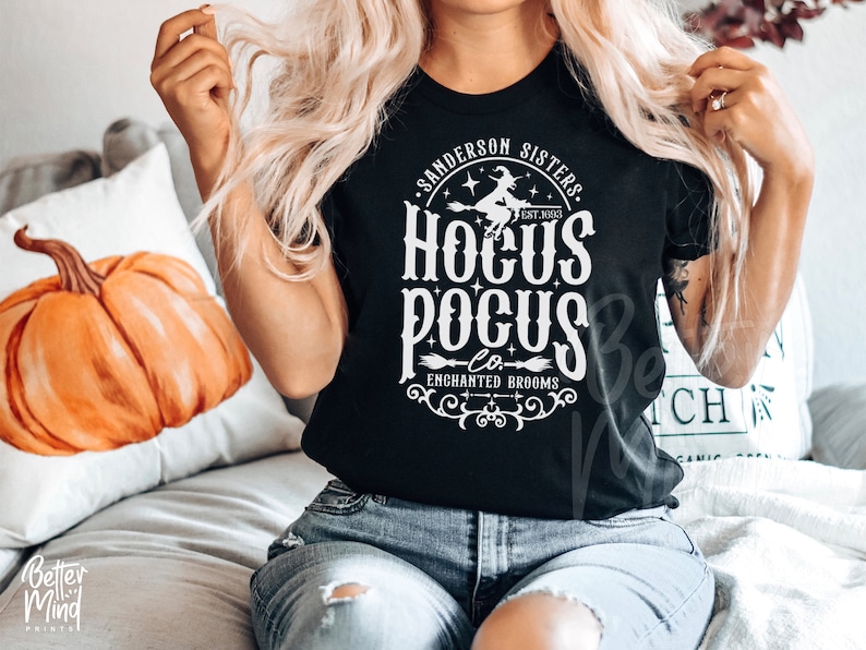 May include: A black short-sleeved t-shirt featuring a white graphic print that reads Sanderson Sisters Est. 1693 Hocus Pocus Co. Enchanted Brooms, accompanied by an illustration of a witch flying on a broomstick, stars, and decorative scrollwork.