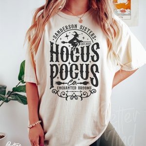 May include: A cream-colored short-sleeve t-shirt featuring a black graphic print that reads Sanderson Sisters Hocus Pocus Co. Enchanted Brooms Est. 1693, accompanied by an illustration of a witch flying on a broomstick and decorative flourishes.