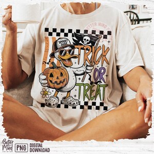 May include: Cream-colored t-shirt featuring a cartoon duck wearing sunglasses and a hat that says "Spooky Dude." The duck holds a carved pumpkin and a flag that says "Trick or Treat." The shirt has a Halloween theme.