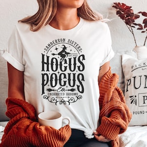 May include: A white t-shirt featuring a black graphic print that reads Sanderson Sisters, Hocus Pocus Co., Enchanted Brooms, and Est. 1693. The design includes a silhouette of a witch flying on a broomstick, stars, and decorative scrollwork.