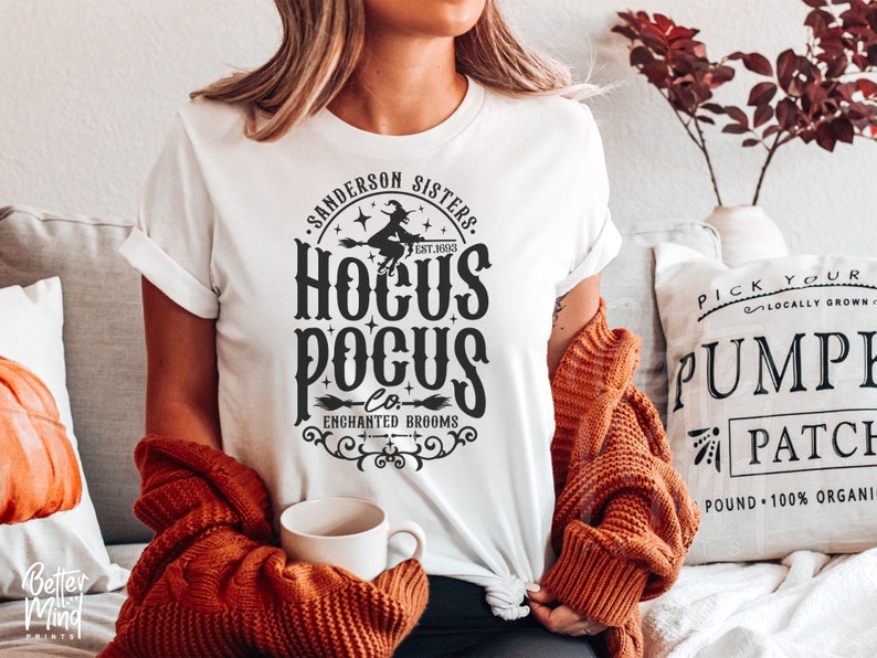 May include: A white t-shirt featuring a black graphic print that reads Sanderson Sisters, Hocus Pocus Co., Enchanted Brooms, and Est. 1693. The design includes a silhouette of a witch flying on a broomstick, stars, and decorative scrollwork.