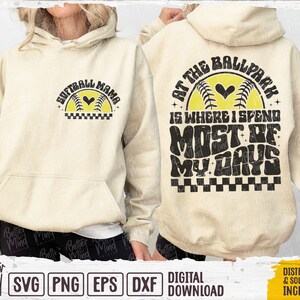May include: A beige hoodie with a graphic design on the front that says "Softball Mama" and a graphic design on the back that says "At the Ballpark is where I spend most of my days".