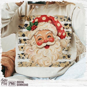 May include: A beige long-sleeve sweatshirt featuring a graphic of Santa Claus with a white beard, red cheeks, and a red polka-dot hat with holly. The design is set against a background of horizontal stripes with a black and tan leopard print pattern.