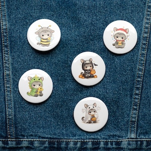 May include: Five white, circular badges with cute animal illustrations in costumes. One shows a bee, another a dragon, a third a cat in a Santa hat, a fourth a bunny in a Halloween costume, and the last a ghost.
