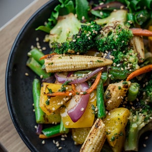 Op de afbeelding: Een zwarte kom gevuld met een kleurrijke roerbak van groenten, waaronder broccoli, maïs, paprika, sperziebonen en rode ui. De groenten zijn gemengd in een lichte saus en bestrooid met sesamzaadjes.