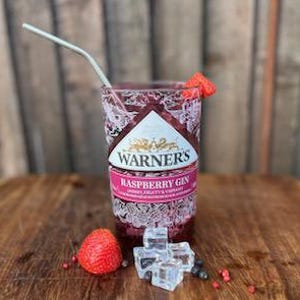 May include: A glass of raspberry gin with ice cubes and a fresh raspberry garnish. The glass has a Warner's Raspberry Gin label with a pink and white design.