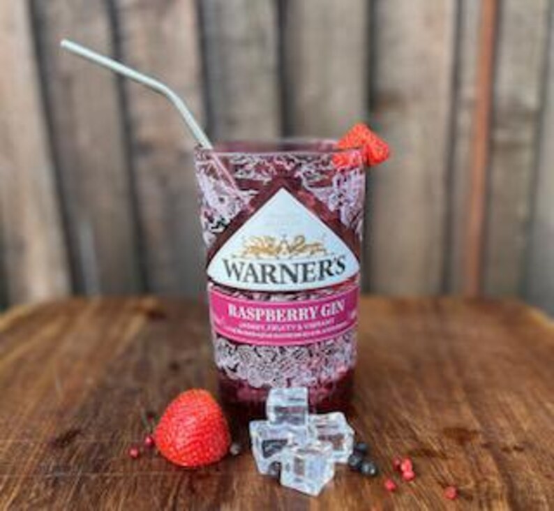 May include: A glass of raspberry gin with ice cubes and a fresh raspberry garnish. The glass has a Warner's Raspberry Gin label with a pink and white design.