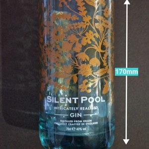 K&ouml;nnte beinhalten: Ein klares Glas, 170 mm hoch und 83 mm breit, mit t&uuml;rkisfarbenem Boden und goldenem Blumendesign. Der Text "SILENT POOL GIN" ist sichtbar. Der Gin wird aus Getreide destilliert und pr&auml;zise in England hergestellt.