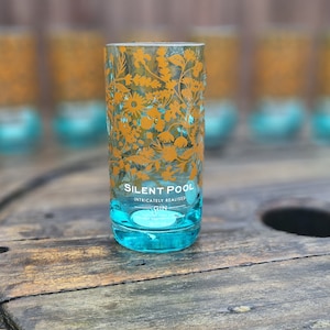 K&ouml;nnte beinhalten: Ein klares, zylindrisches Glas mit t&uuml;rkisfarbenem Boden und goldenem floralem Design. Auf dem Glas steht "SILENT POOL" und "INTRICATELY REALISED GIN". Mehrere &auml;hnliche Gl&auml;ser sind im Hintergrund unscharf.