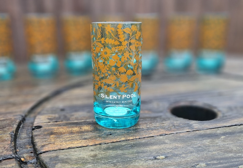 K&ouml;nnte beinhalten: Ein klares, zylindrisches Glas mit t&uuml;rkisfarbenem Boden und goldenem floralem Design. Auf dem Glas steht "SILENT POOL" und "INTRICATELY REALISED GIN". Mehrere &auml;hnliche Gl&auml;ser sind im Hintergrund unscharf.