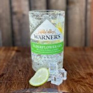 May include: A clear glass bottle of Warner's Elderflower Gin with a lime wedge and ice cubes on a wooden surface.