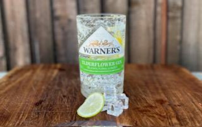May include: A clear glass bottle of Warner's Elderflower Gin with a lime wedge and ice cubes on a wooden surface.