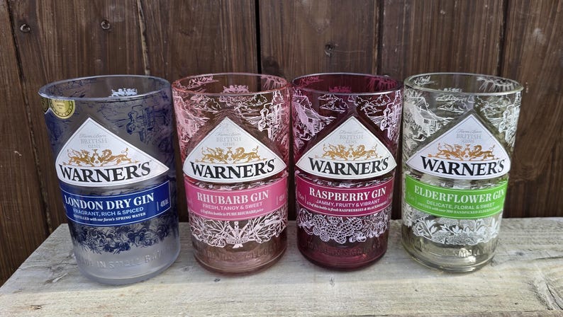 May include: Four glass bottles with different colored labels and Warner's Gin printed on them. The labels are for London Dry Gin, Rhubarb Gin, Raspberry Gin, and Elderflower Gin.