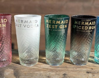 May include: Five tall, textured glasses with the words "Mermaid" and various spirit names printed in gold. The glasses are colored pink, white, green, black, and blue. The glasses are displayed on a wooden surface.