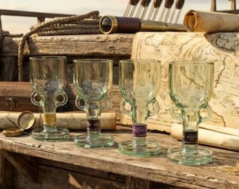 May include: Four clear glass goblets with a unique design, featuring small handles and a wide base. The goblets are displayed on a rustic wooden surface, with nautical-themed props in the background, including a map and a telescope.
