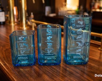 May include: Three blue square-shaped glass vases of varying heights, arranged on a wooden surface. Each vase features botanical illustrations and text, including "Hand Selected Botanicals." The vases are in a bar setting.