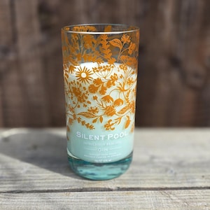 K&ouml;nnte beinhalten: Eine hohe Glaskerze mit einem floralen Design in Orange. Die Kerze hat einen hellblauen Sockel und eine wei&szlig;e Kerze im Inneren. Der Text "SILENT POOL" ist auf dem Glas sichtbar. Die Kerze steht auf einer Holzoberfl&auml;che.