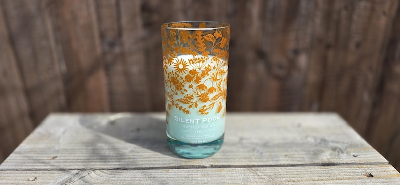 K&ouml;nnte beinhalten: Eine hohe Glaskerze mit einem floralen Design in Orange. Die Kerze hat einen hellblauen Sockel und eine wei&szlig;e Kerze im Inneren. Der Text "SILENT POOL" ist auf dem Glas sichtbar. Die Kerze steht auf einer Holzoberfl&auml;che.