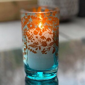 K&ouml;nnte beinhalten: Eine brennende Kerze in einem t&uuml;rkisfarbenen Glas mit orangefarbenen floralen Mustern. Die Kerze hat einen wei&szlig;en Innenraum und der Text "SILENT POOL" ist sichtbar. Das Glas ist zylindrisch.