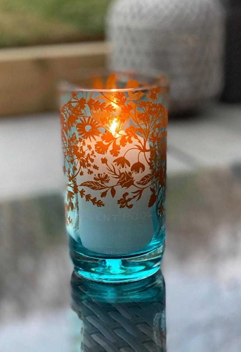 K&ouml;nnte beinhalten: Eine brennende Kerze in einem t&uuml;rkisfarbenen Glas mit orangefarbenen floralen Mustern. Die Kerze hat einen wei&szlig;en Innenraum und der Text "SILENT POOL" ist sichtbar. Das Glas ist zylindrisch.