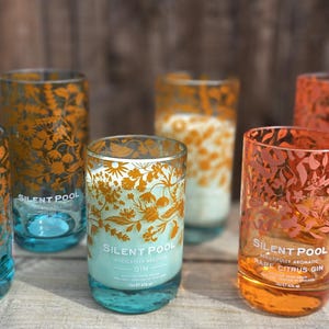 K&ouml;nnte beinhalten: Sechs klare, zylindrische Gl&auml;ser mit floralen Designs in Orange- und Goldt&ouml;nen. Die Gl&auml;ser haben einen t&uuml;rkisblauen Boden und der Text "SILENT POOL" ist auf einigen sichtbar. Die Gl&auml;ser sind auf einer Holzoberfl&auml;che angeordnet.