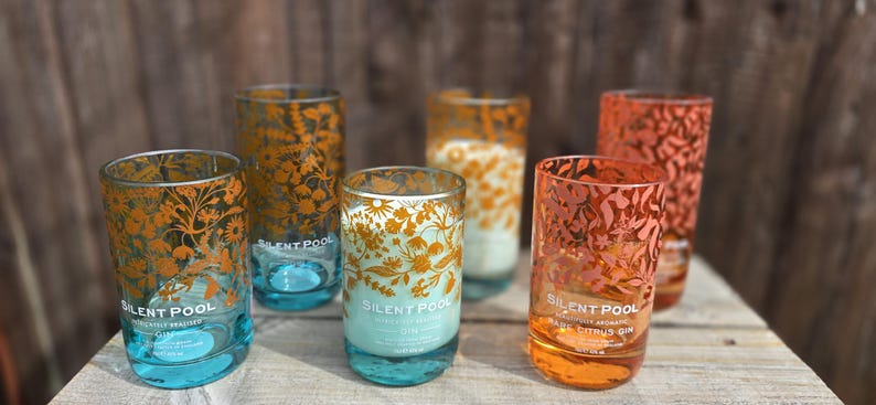 K&ouml;nnte beinhalten: Sechs klare, zylindrische Gl&auml;ser mit floralen Designs in Orange- und Goldt&ouml;nen. Die Gl&auml;ser haben einen t&uuml;rkisblauen Boden und der Text "SILENT POOL" ist auf einigen sichtbar. Die Gl&auml;ser sind auf einer Holzoberfl&auml;che angeordnet.