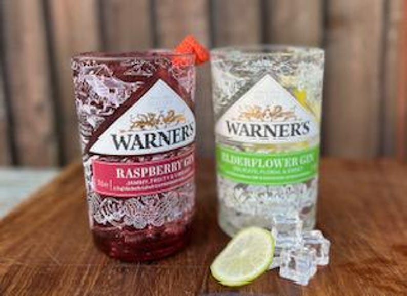 May include: Two clear glass bottles with Warner's Gin labels. The bottle on the left is raspberry gin and the bottle on the right is elderflower gin. Both bottles are filled with ice and garnished with fruit.