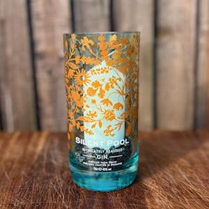 K&ouml;nnte beinhalten: Ein zylindrisches Glas mit t&uuml;rkisfarbenem Boden und einem floralen Design in Orange und Wei&szlig;. Der Text "SILENT POOL" und "GIN" sind sichtbar. Das Glas steht auf einer Holzoberfl&auml;che mit verschwommenem Holz-Hintergrund.