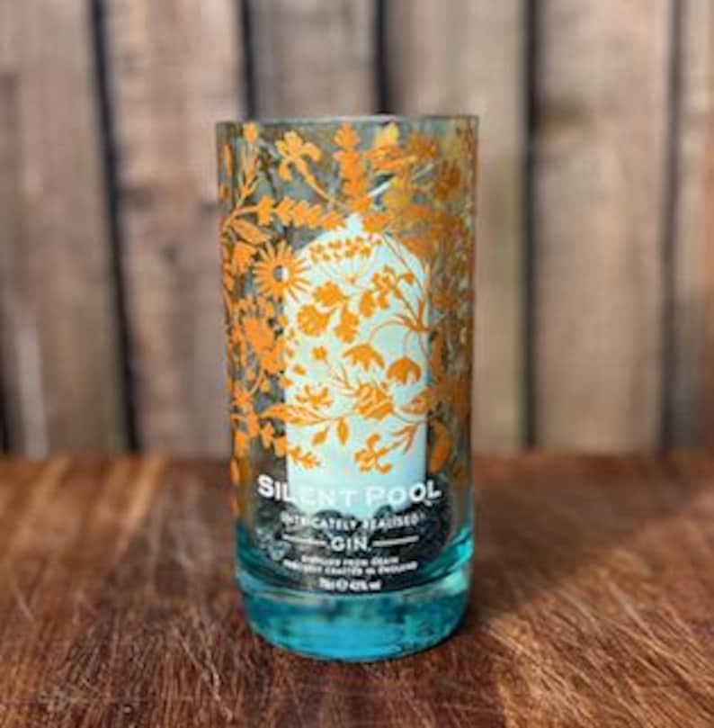 K&ouml;nnte beinhalten: Ein zylindrisches Glas mit t&uuml;rkisfarbenem Boden und einem floralen Design in Orange und Wei&szlig;. Der Text "SILENT POOL" und "GIN" sind sichtbar. Das Glas steht auf einer Holzoberfl&auml;che mit verschwommenem Holz-Hintergrund.