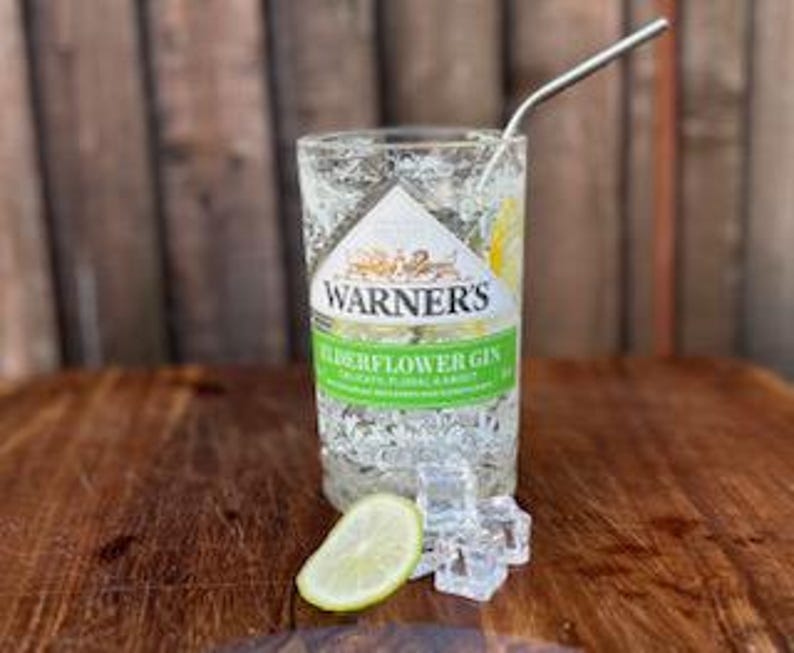 May include: A glass filled with ice and a clear liquid, garnished with a lime wedge. A metal straw is in the glass. The glass has a label that reads "Warner's Elderflower Gin".
