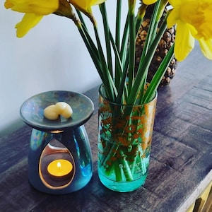 K&ouml;nnte beinhalten: Ein tealfarbener Diffusor f&uuml;r &auml;therische &Ouml;le mit einer brennenden Kerze und zwei wei&szlig;en Wachsschmelzen. Eine klare Glasvase mit floralem Design h&auml;lt Narzissen. Die Gegenst&auml;nde stehen auf einer Holzoberfl&auml;che.