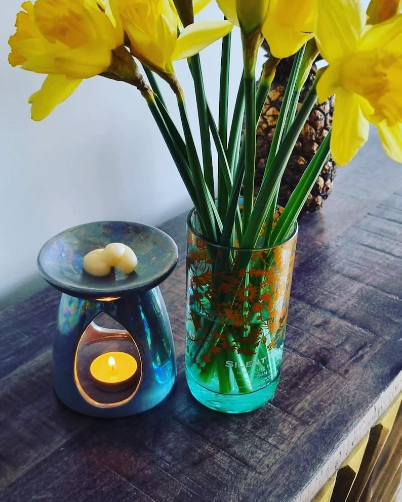 K&ouml;nnte beinhalten: Ein tealfarbener Diffusor f&uuml;r &auml;therische &Ouml;le mit einer brennenden Kerze und zwei wei&szlig;en Wachsschmelzen. Eine klare Glasvase mit floralem Design h&auml;lt Narzissen. Die Gegenst&auml;nde stehen auf einer Holzoberfl&auml;che.