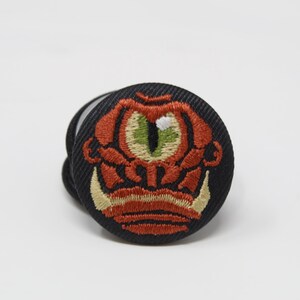 Op de afbeelding: Een ronde zwarte stoffen knoop met een geborduurd ontwerp van een wezen met een groot groen oog, oranje gelaatstrekken en twee opvallende crèmekleurige hoektanden. Het borduurwerk is voorzien van gedetailleerde steken op een donkere ondergrond.