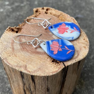 May include: A pair of teardrop-shaped earrings with a blue and white marbled background and a red floral design. The earrings have silver-colored, square-shaped hooks. The earrings are displayed on a wooden surface.