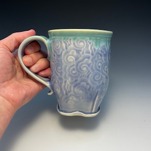 May include: A ceramic mug with a light blue glaze and a white swirl design. The mug has a handle and a rounded bottom.
