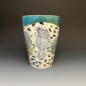 May include: A ceramic tumbler featuring a teal glazed rim and a cream-colored body decorated with black line art of a bird and floral motifs. The design includes scattered blue and grey leaf-shaped accents, creating a detailed pattern across the surface.