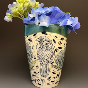 May include: A ceramic vase with a tapered shape, featuring a cream-colored body decorated with black line drawings of a bird and floral patterns. The rim is glazed in a deep teal, and the vase is filled with blue and yellow hydrangea flowers.