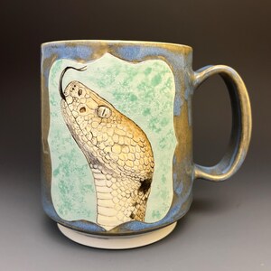 May include: A ceramic mug with a blue and brown mottled glaze featuring a central rectangular panel with a light green textured background. The panel displays a detailed illustration of a snake head in profile with scales and a protruding tongue.